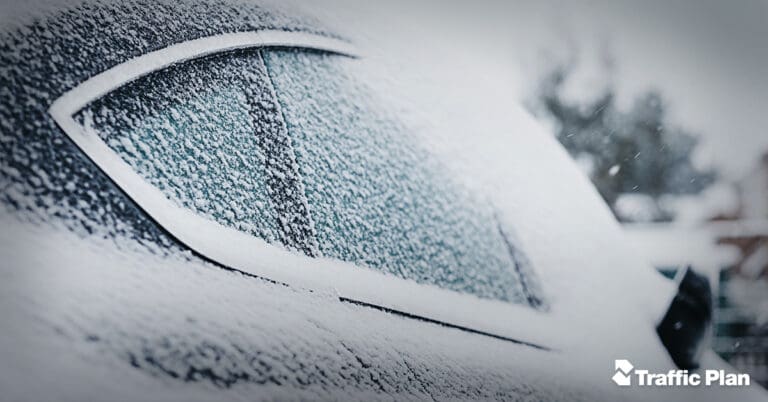 A car covered in snow, with a close-up of the rear window partially obscured by frost. The background is blurred, and the words Traffic Plan appear in the bottom right corner.