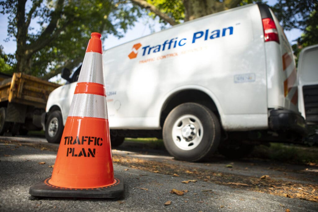 A bright orange traffic cone labeled TRAFFIC PLAN stands on pavement in front of a white van with the Traffic Plan logo, parked near trees and other vehicles on a sunny day.