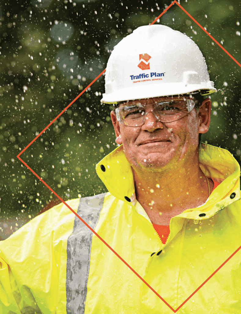 A construction worker in a yellow high-visibility jacket and white hard hat stands outdoors in the rain. The hard hat has a Traffic Plan logo, and water droplets are captured midair around the person.