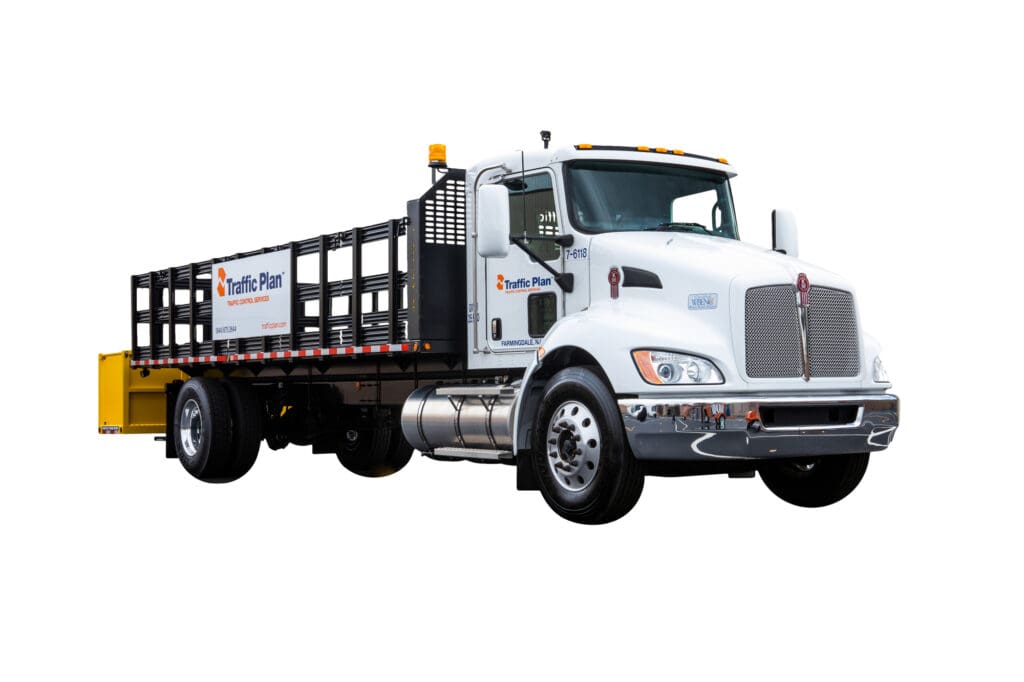 A white flatbed truck with “Traffic Plan” logos on the door and flatbed side, featuring a metal railing bed and yellow equipment at the rear, isolated on a white background.