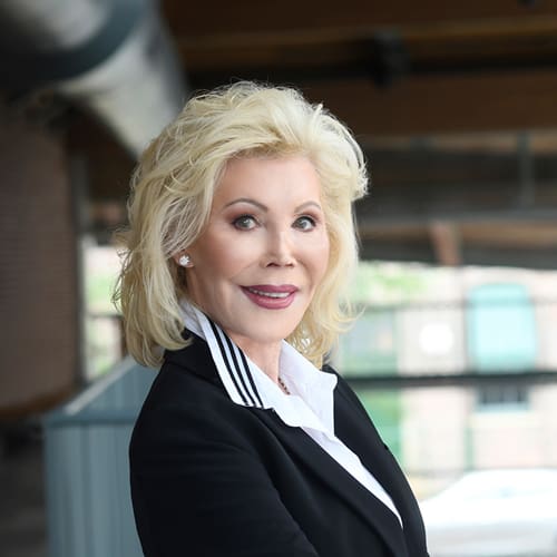 A smiling woman with shoulder-length blond hair, wearing a black blazer with white trim and a white shirt, stands in front of a blurred urban background.