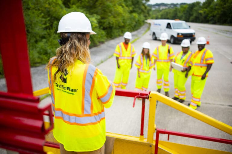 A group of six workers in high-visibility clothing and hard hats stand on a road, listening to a colleague who is elevated above them, with greenery and a work vehicle in the background.