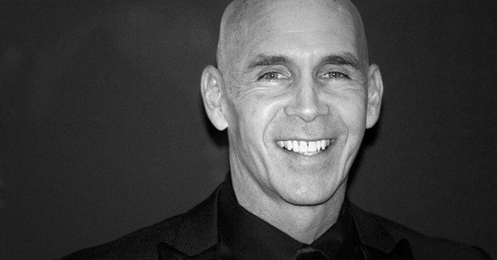 A bald man in a suit and tie smiles warmly at the camera against a dark, plain background. The image is in black and white.