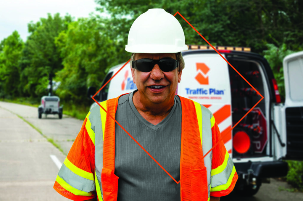 A man wearing a white hard hat, sunglasses, and a reflective safety vest stands smiling in front of a Traffic Plan work van parked on the side of a road with trees in the background.