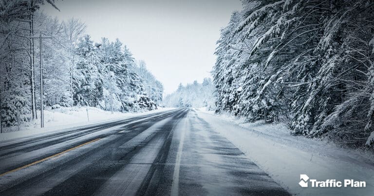 A two-lane road runs through a snowy landscape with trees covered in snow on both sides under an overcast sky. Construction cones line the roadside, hinting at ongoing work. The scene appears cold and wintry. Traffic Plan logo is in the bottom right corner.