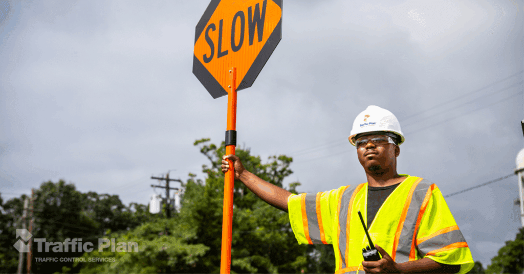 A flagger in a hard hat and reflective vest holds a SLOW sign and a walkie-talkie, standing outdoors near trees and power lines. The Traffic Plan logo appears in the bottom left corner.