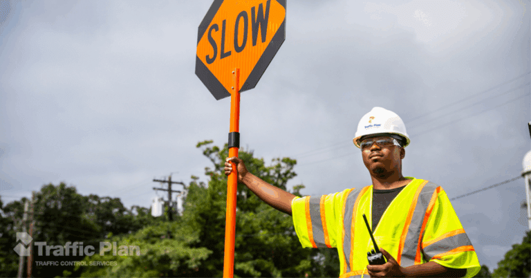 A flagger in a hard hat and reflective vest holds a SLOW sign and a walkie-talkie, standing outdoors near trees and power lines. The Traffic Plan logo appears in the bottom left corner.