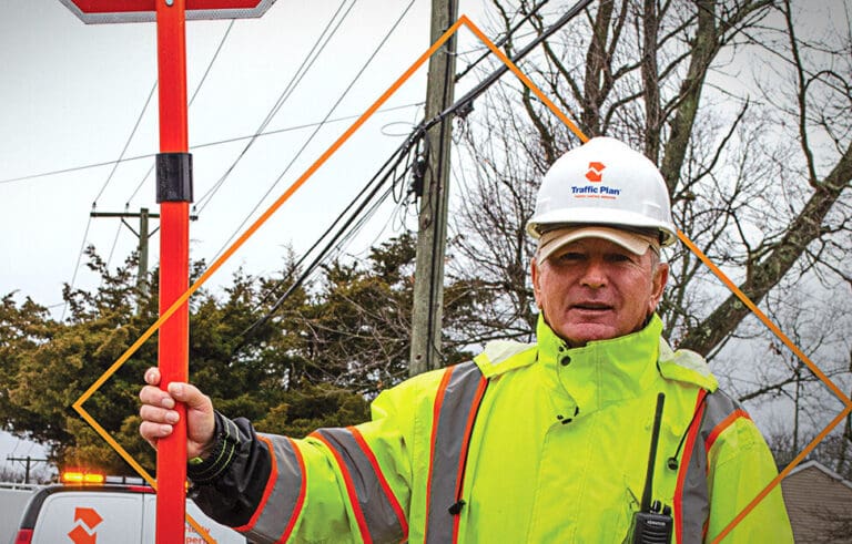 Employee Spotlight: A construction worker wearing a neon yellow safety jacket and white hard hat stands outdoors, holding a red sign. Trees, power lines, and a utility truck are visible in the background.