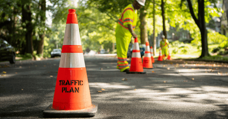 A row of orange traffic cones labeled TRAFFIC PLAN lines a sunlit street, while a worker in a reflective safety vest and helmet arranges cones in the background—highlighting organized traffic management services amid lush greenery.