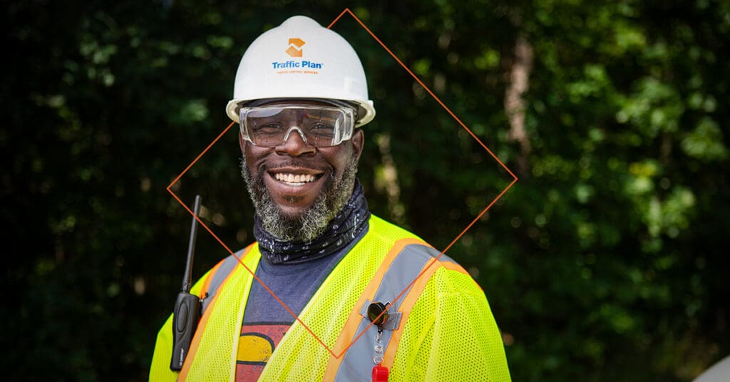 A smiling construction worker wearing a white hard hat, safety glasses, and a neon yellow safety vest stands outdoors with greenery in the background. A walkie-talkie is clipped to the vest.