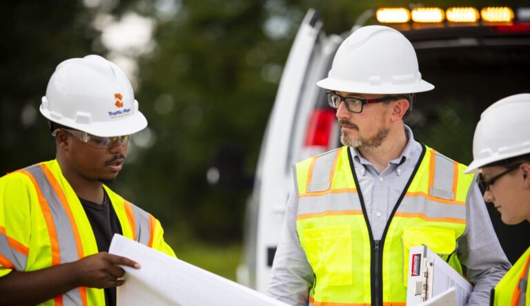 Three construction workers wearing safety vests, hard hats, and glasses discuss blueprints outdoors near a work vehicle. One holds the blueprints, while another holds a clipboard. Trees are visible in the background.