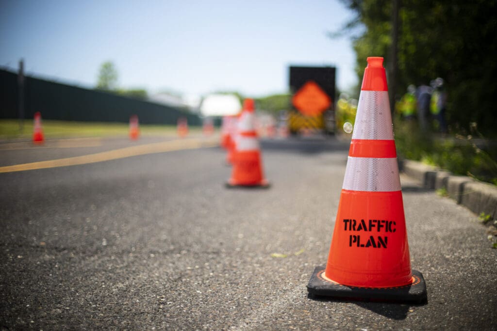 Bright orange traffic cones line a paved road, with the nearest cone labeled TRAFFIC PLAN. In the background, workers in safety vests handle roadwork near greenery—a typical scene for those seeking traffic plan jobs.