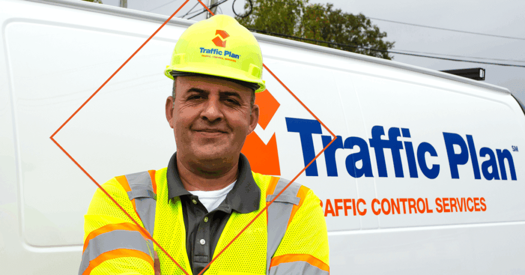 A man wearing a fluorescent safety vest and hard hat stands in front of a white van labeled Traffic Plan Traffic Control Services. He is smiling with his arms crossed.