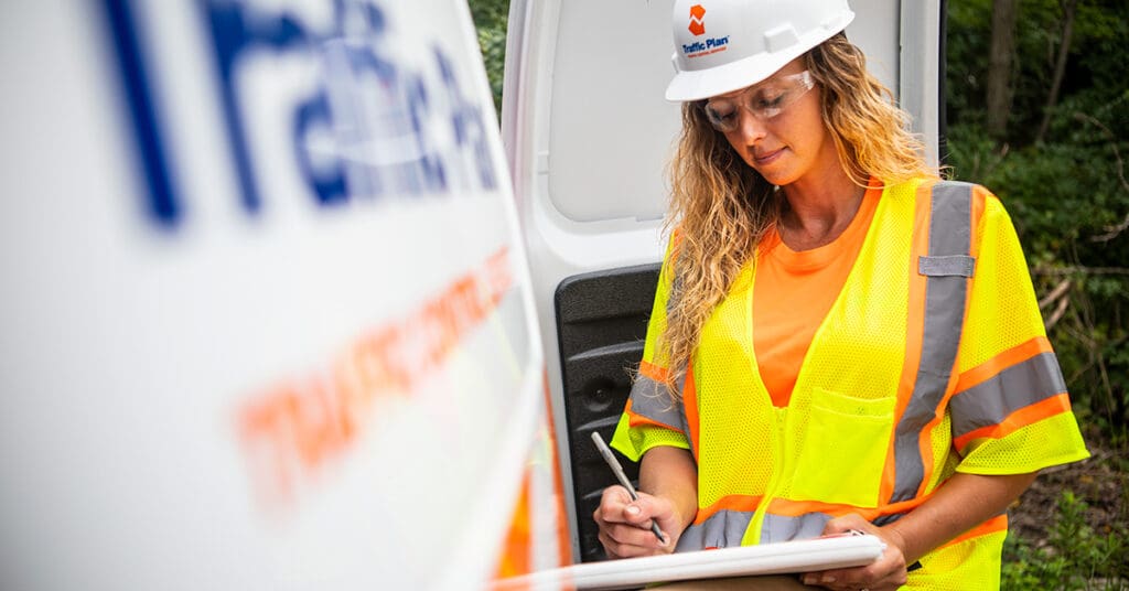 A woman in a hard hat, safety glasses, and a reflective yellow vest writes on a clipboard outdoors next to a white vehicle.