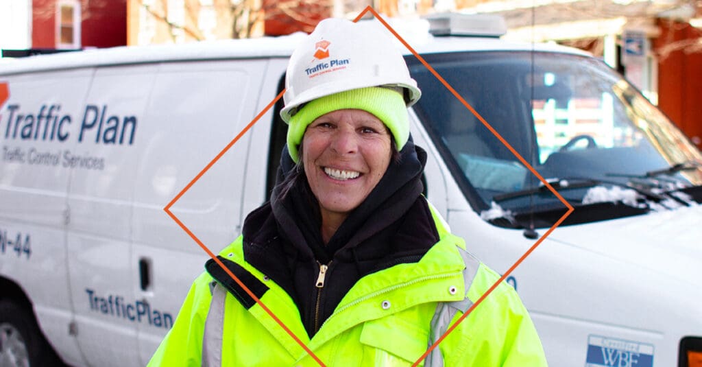 A smiling construction worker wearing a neon yellow jacket and hard hat stands in front of a white Traffic Plan van on a snowy street.