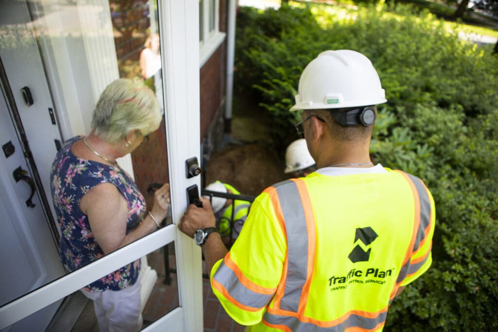A worker wearing a helmet and a Traffic Plan safety vest stands by a doorway, while an older woman with short gray hair and floral top talks to him. Another worker in safety gear is visible near bushes outside.