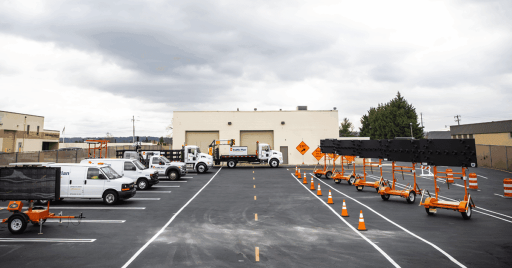 A parking lot with several white utility trucks, orange traffic cones, and electronic road signs on trailers, in front of a beige industrial building under a cloudy sky.