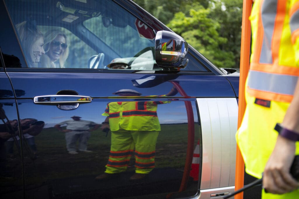 A woman smiles inside a parked car near a worker in a neon yellow safety vest. The worker’s reflection is visible on the car door, with trees and grass in the background.