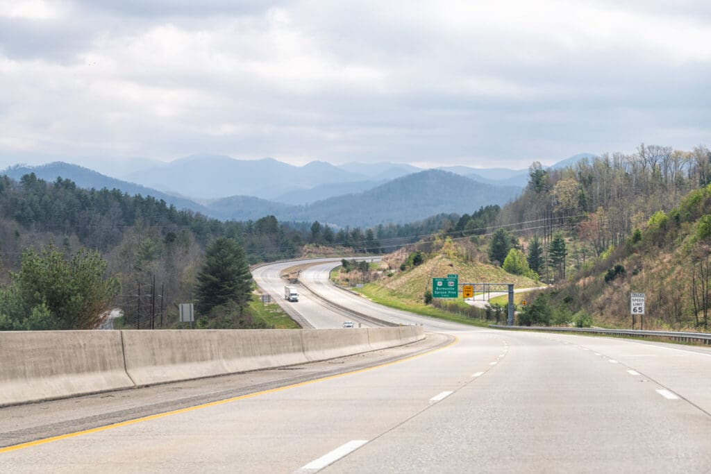 A mostly empty highway curves through a scenic landscape of rolling hills and mountains under a cloudy sky, with trees lining both sides and road signs visible along the roadside.