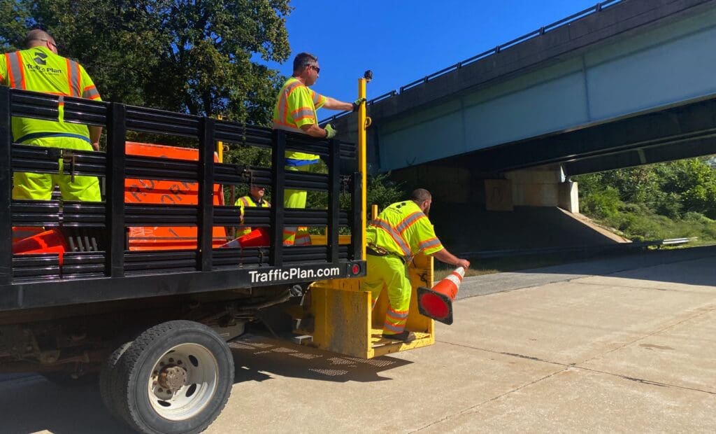 Several road workers in neon safety uniforms unload orange traffic cones from a black truck marked TrafficPlan.com near an overpass on a sunny day.