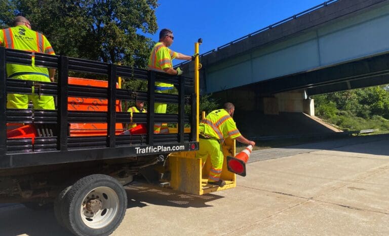 Several road workers in neon safety uniforms unload orange traffic cones from a black truck marked TrafficPlan.com near an overpass on a sunny day.