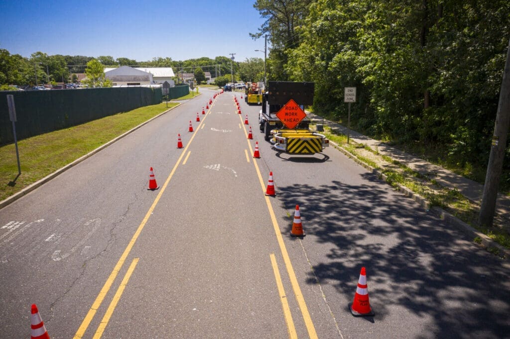 A two-lane road lined with orange traffic cones, narrowing into one lane for road work. A construction sign reads ROAD WORK AHEAD, and trees and buildings are visible along the sides.