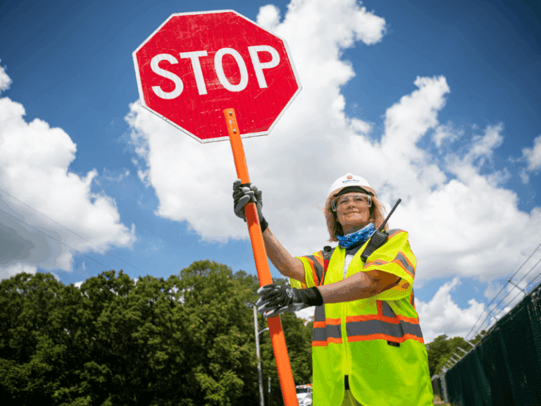 A construction worker wearing a safety vest, hard hat, and gloves holds a large red stop sign. The person is standing outdoors on a sunny day, with trees and a blue sky with clouds in the background.