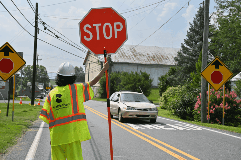 A construction worker in a fluorescent vest and hard hat holds a stop sign, directing traffic as a car waits on a rural road lined with warning signs and greenery.