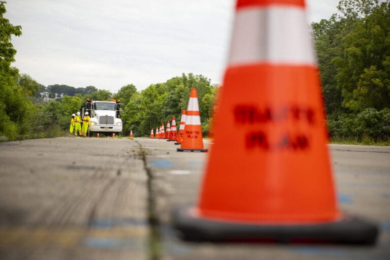 A row of orange traffic cones lines a road, with a work truck and several workers in yellow safety vests visible in the background surrounded by greenery. The focus is on the cones in the foreground.