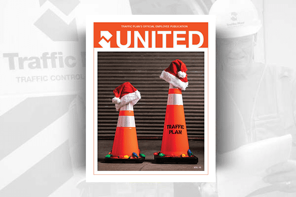 Cover of United magazine featuring two orange traffic cones wearing Santa hats, with holiday lights at their bases. The cones are labeled Traffic Plan. A blurred worker and company logo appear in the background.