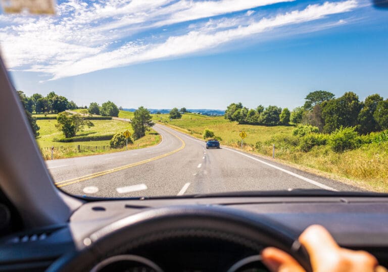 View from inside a car driving on a winding rural road, with one hand on the steering wheel. The road is surrounded by green fields, trees, and a blue sky with wispy clouds. A car is ahead in the distance.