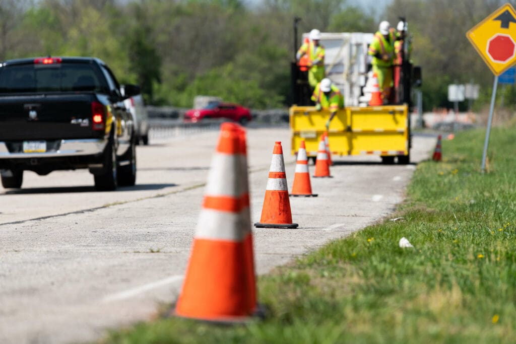 Several orange traffic cones mark off a road lane where construction workers in safety vests and helmets work near a yellow truck. Vehicles pass by on the left, and trees are visible in the background.