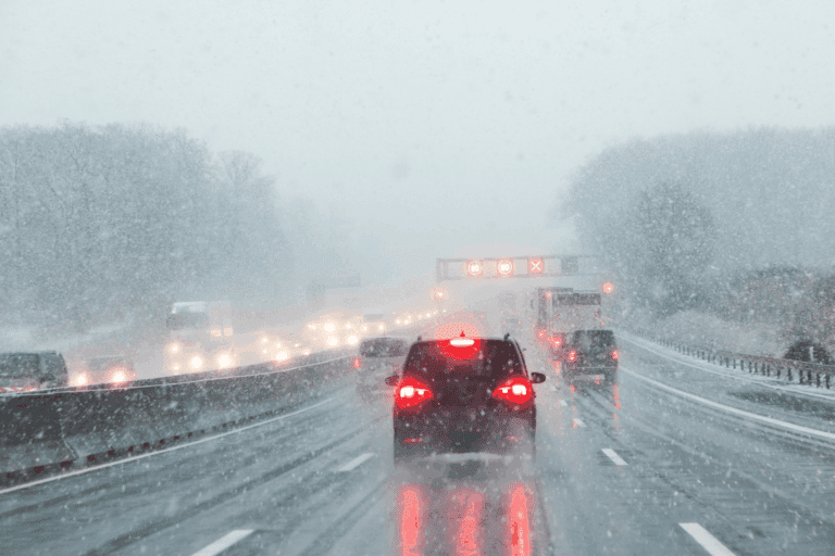 Cars driving on a wet highway during heavy snowfall, with low visibility, blurry taillights, and road signs visible in the distance. Trees line the roadside, and everything is covered in snow.