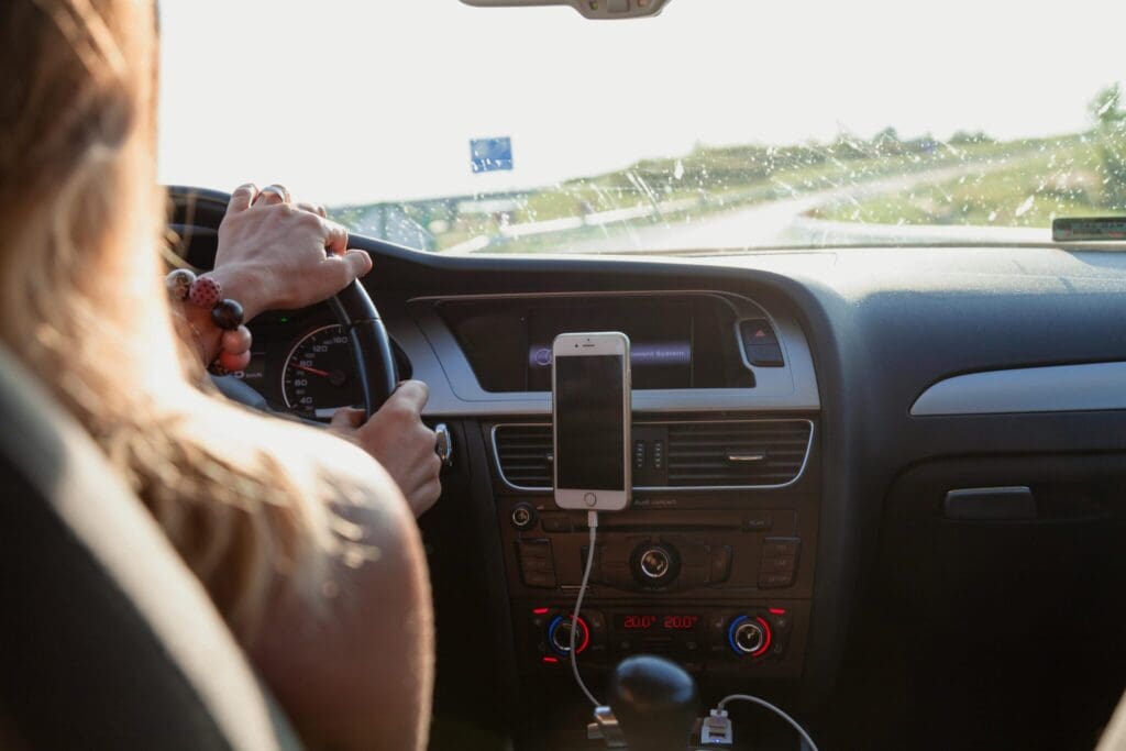 A person drives a car during the day with both hands on the steering wheel. A smartphone is mounted on the dashboard and connected to a charging cable. Sunlight shines through the windshield.