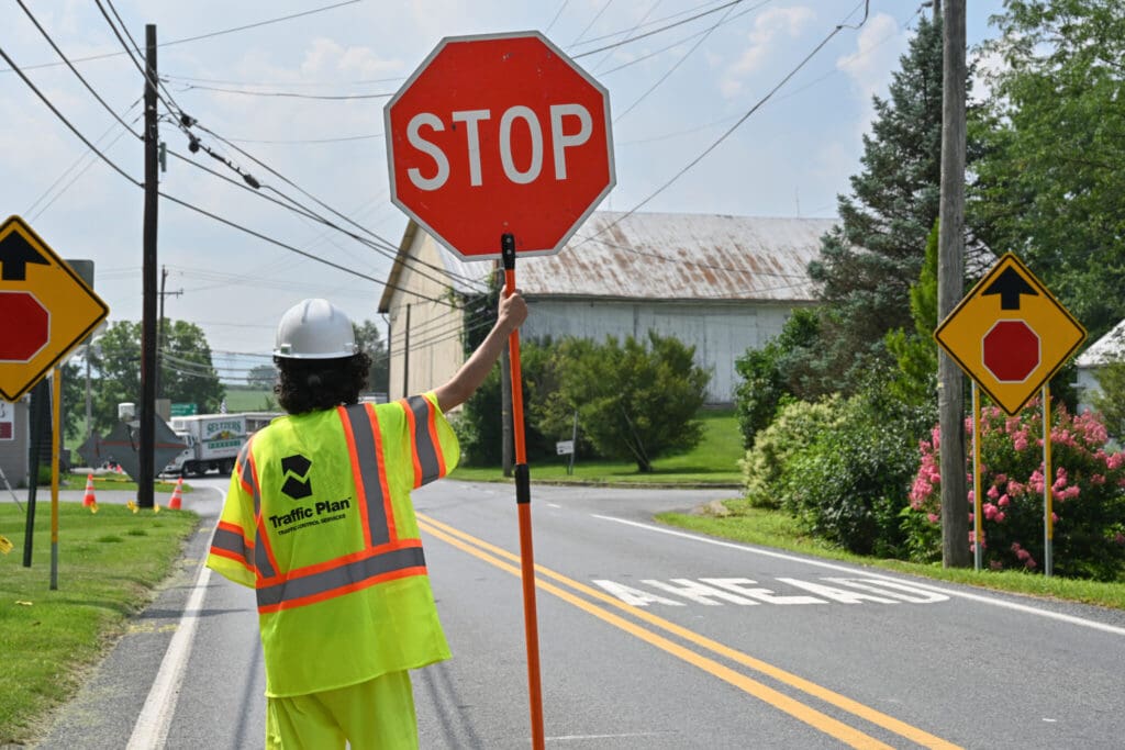 A construction worker in a neon yellow vest and white hard hat holds a “STOP” sign by a rural road, with traffic signs and a barn visible in the background.