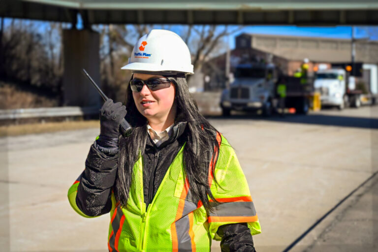 A construction worker in a hard hat, safety vest, and sunglasses holds a walkie-talkie on a sunny day. Trucks and equipment are visible on the road behind her, under an overpass.