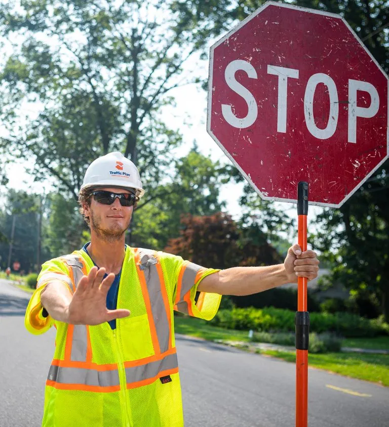 A construction worker wearing a hard hat, sunglasses, and a neon safety vest holds a large stop sign and raises his hand to signal traffic to stop on a sunny day.