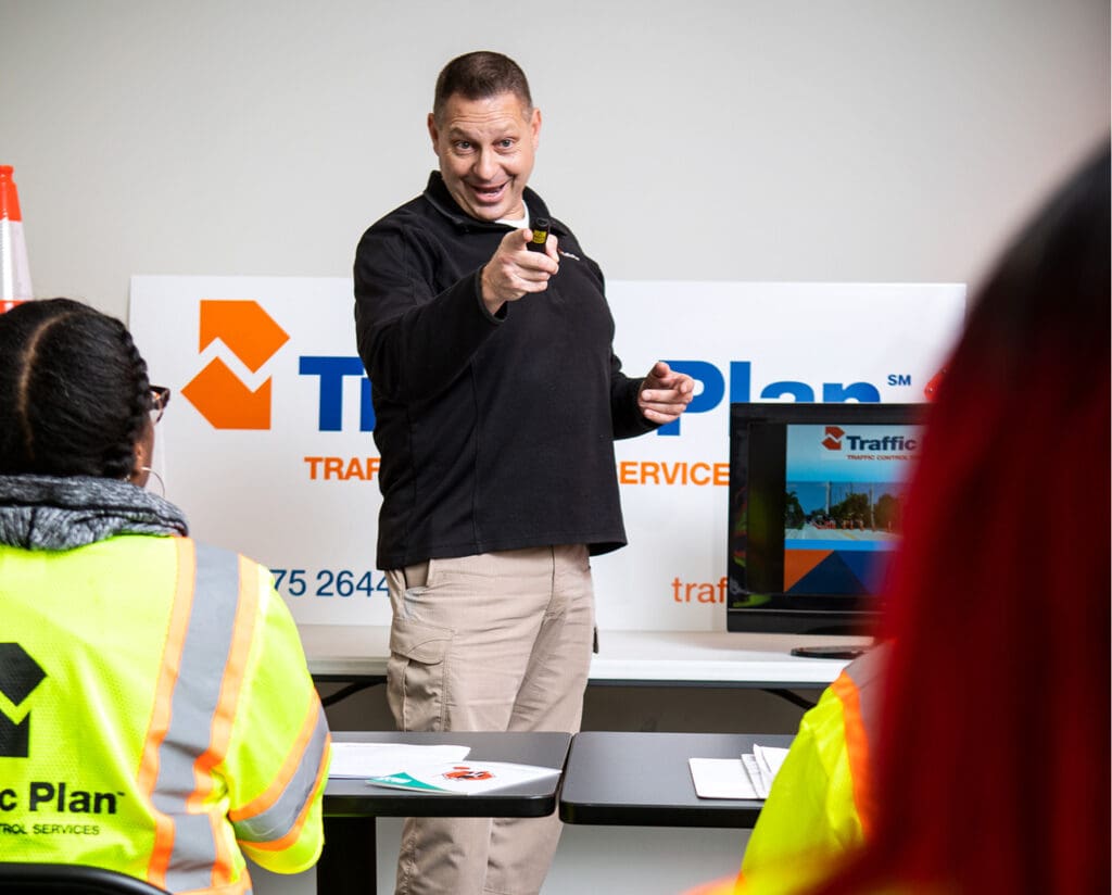 A man in a black shirt points and smiles while giving a presentation to people wearing yellow safety vests at a training session. A “Traffic Plan” sign and a monitor are visible in the background.