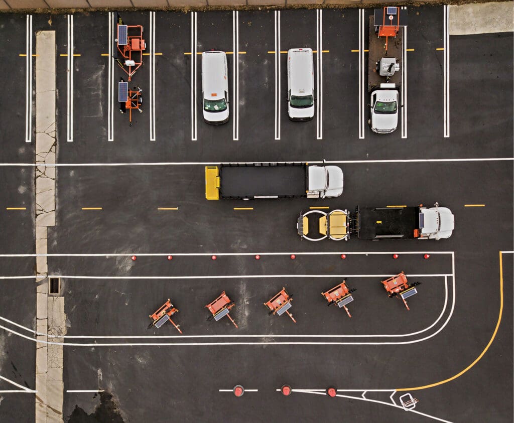Aerial view of a parking lot with several parked vehicles, including cars and trucks, as well as orange roadwork equipment lined up and marked parking spaces, with visible road markings and traffic cones.