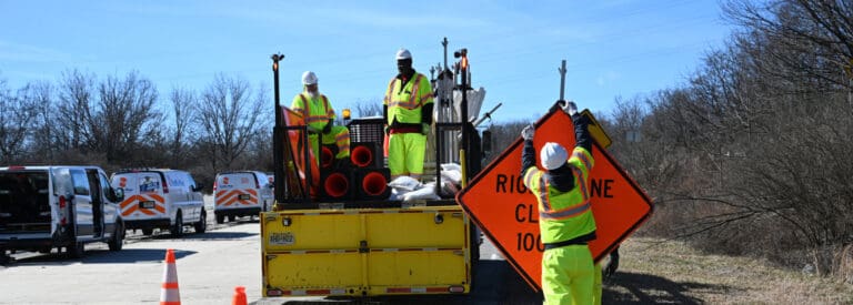 Three road workers in high-visibility gear set up a Right Lane Closed sign from a yellow utility truck beside a rural road, with work vans and traffic cones nearby. Leafless trees line the background.
