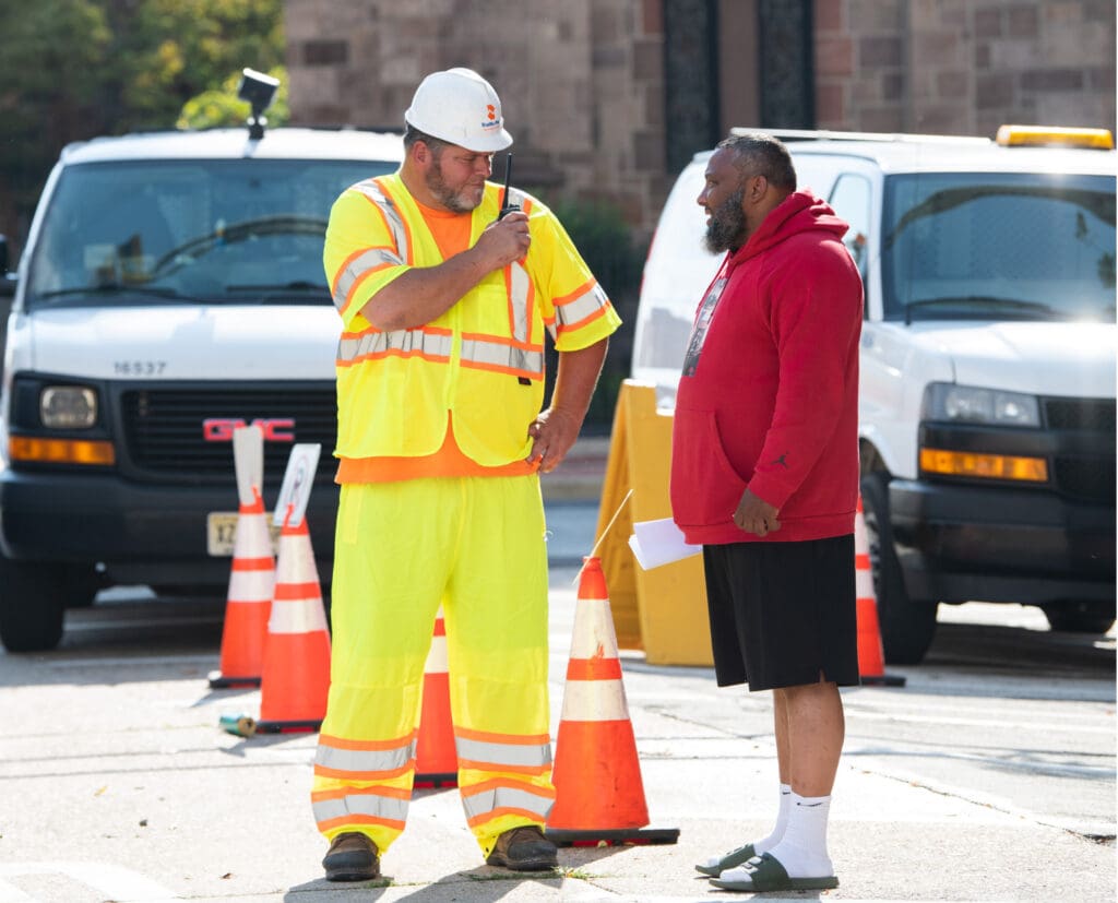 A construction worker in a fluorescent yellow safety suit and hard hat talks on a radio while facing a man in a red hoodie and black shorts. Traffic cones and white vans are in the background.