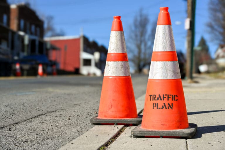 Two orange traffic cones stand on the edge of a sidewalk next to a street, with one cone labeled TRAFFIC PLAN. Blurred buildings and more cones are visible in the background.