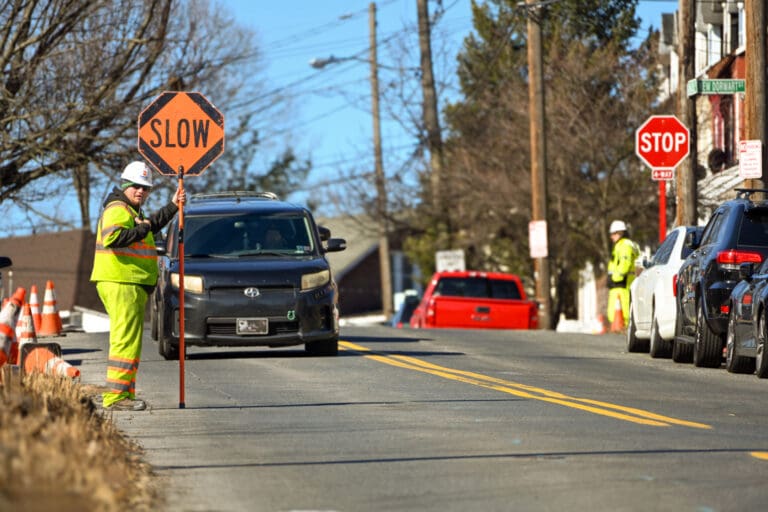 A construction worker in a safety vest and helmet holds a SLOW sign to direct traffic on a sunny street with cones and cars. Another worker stands near a stop sign farther down the road.