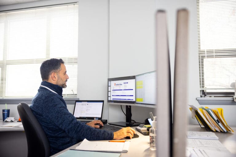 A man sits at a desk in an office, working on a computer with two monitors and a laptop. Papers, folders, and a cup are on the desk. Sunlight streams in through the windows behind him.