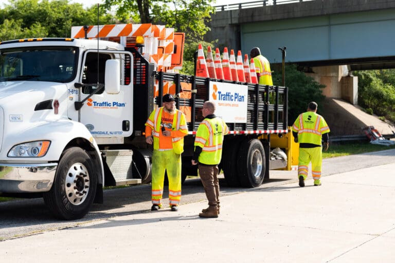 Four road workers in high-visibility clothing stand near a Traffic Plan truck loaded with orange traffic cones on the side of a road, with a bridge and greenery in the background.