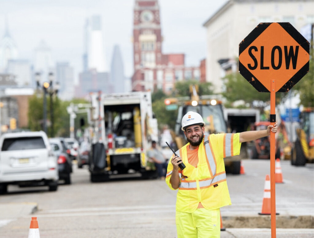 A construction worker in a neon safety uniform and hard hat holds a SLOW sign and a walkie-talkie on a city street with traffic, equipment, and buildings in the background.