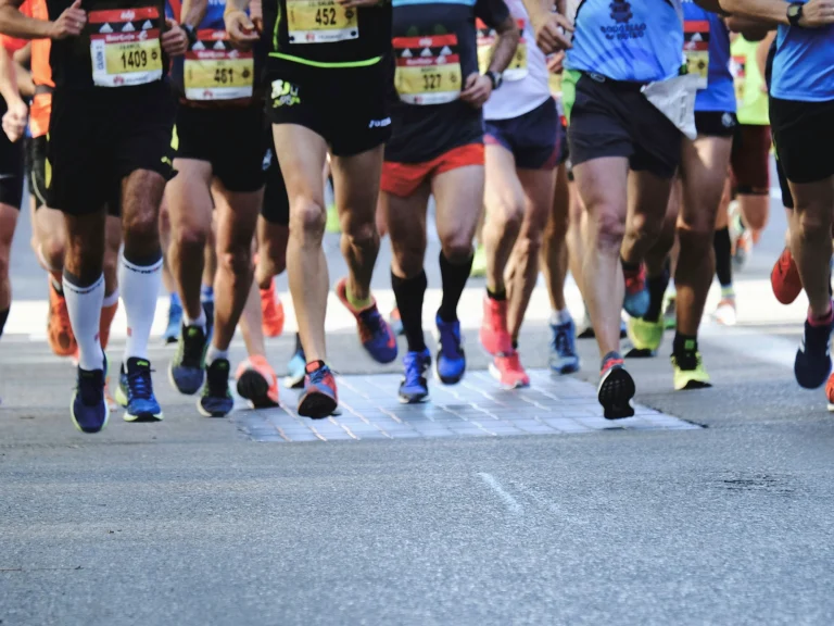 A group of runners in athletic gear participate in a road race, with only their legs and running shoes visible as they move forward on a paved street.