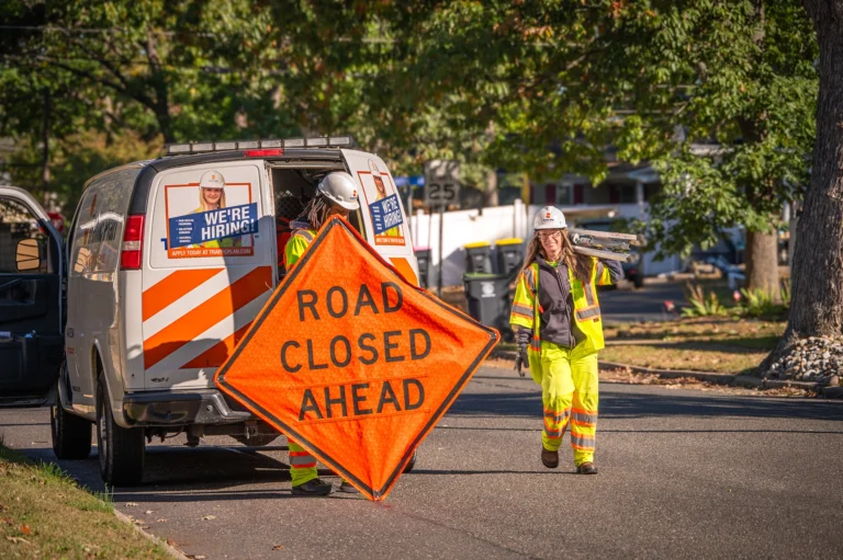 Two utility workers in neon safety gear set up a bright orange Road Closed Ahead sign near a work van with a Were Hiring! sign on its door, on a sunny residential street.