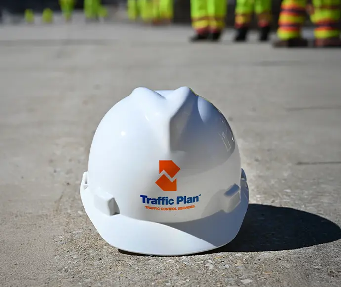 A white hard hat with the Traffic Plan logo sits on pavement, with workers in high-visibility clothing blurred in the background.