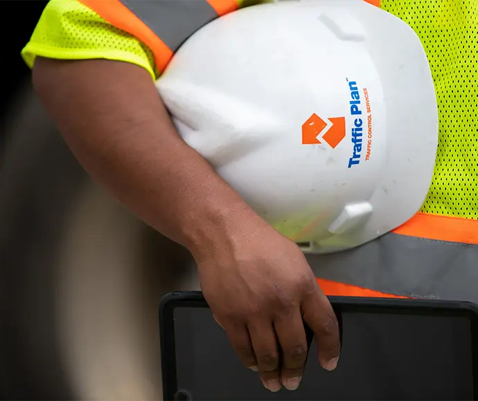 A person wearing a yellow safety vest with orange stripes holds a white hard hat labeled Traffic Plan and a black tablet.